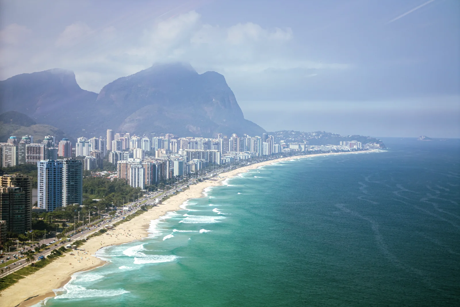 Orla da Barra da Tijuca vista do alto entre a cidade, o oceano e as montanhas.
